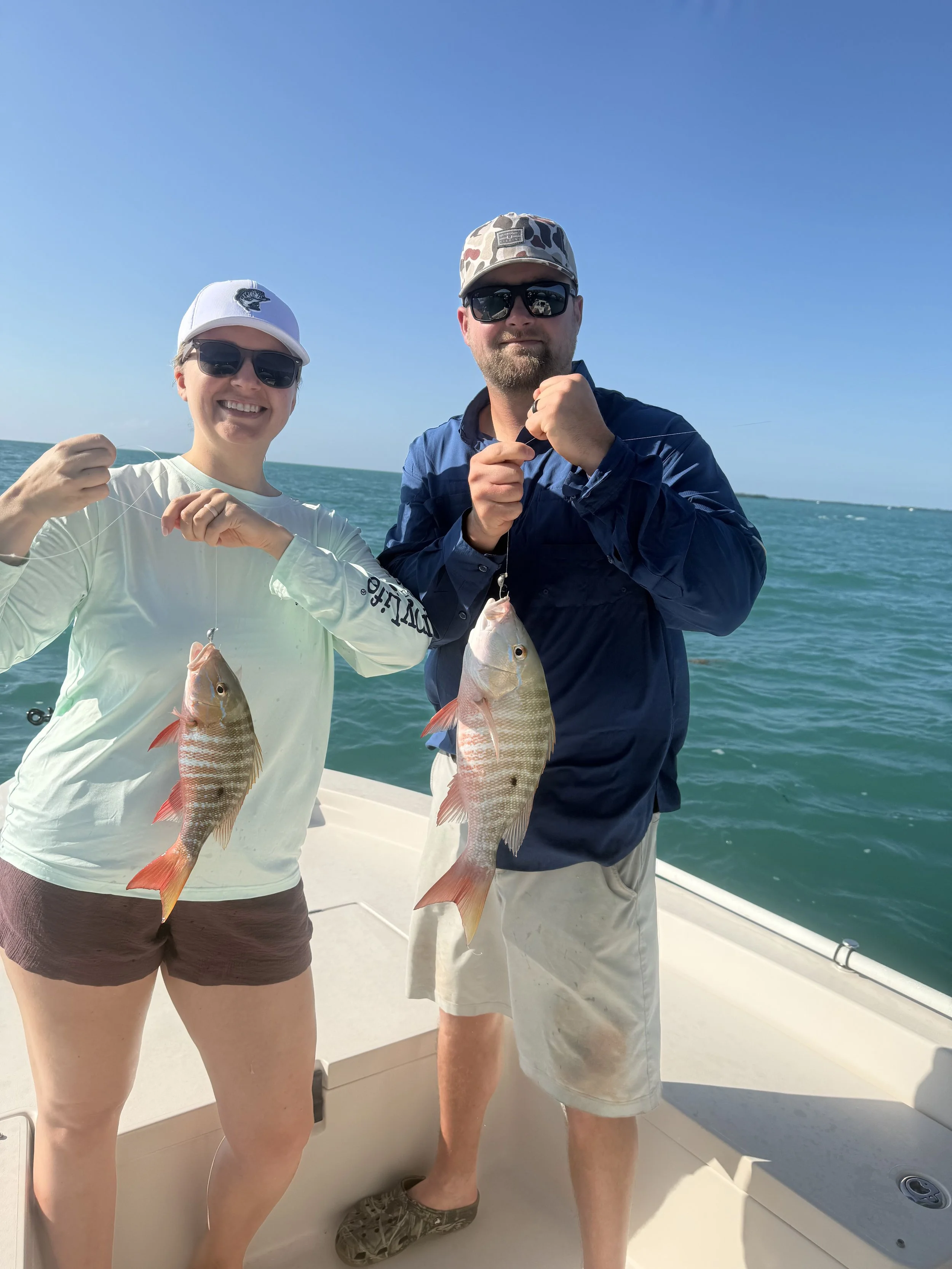 A smiling woman and man on a boat holding up two fish they caught, with the ocean and blue sky in the background.