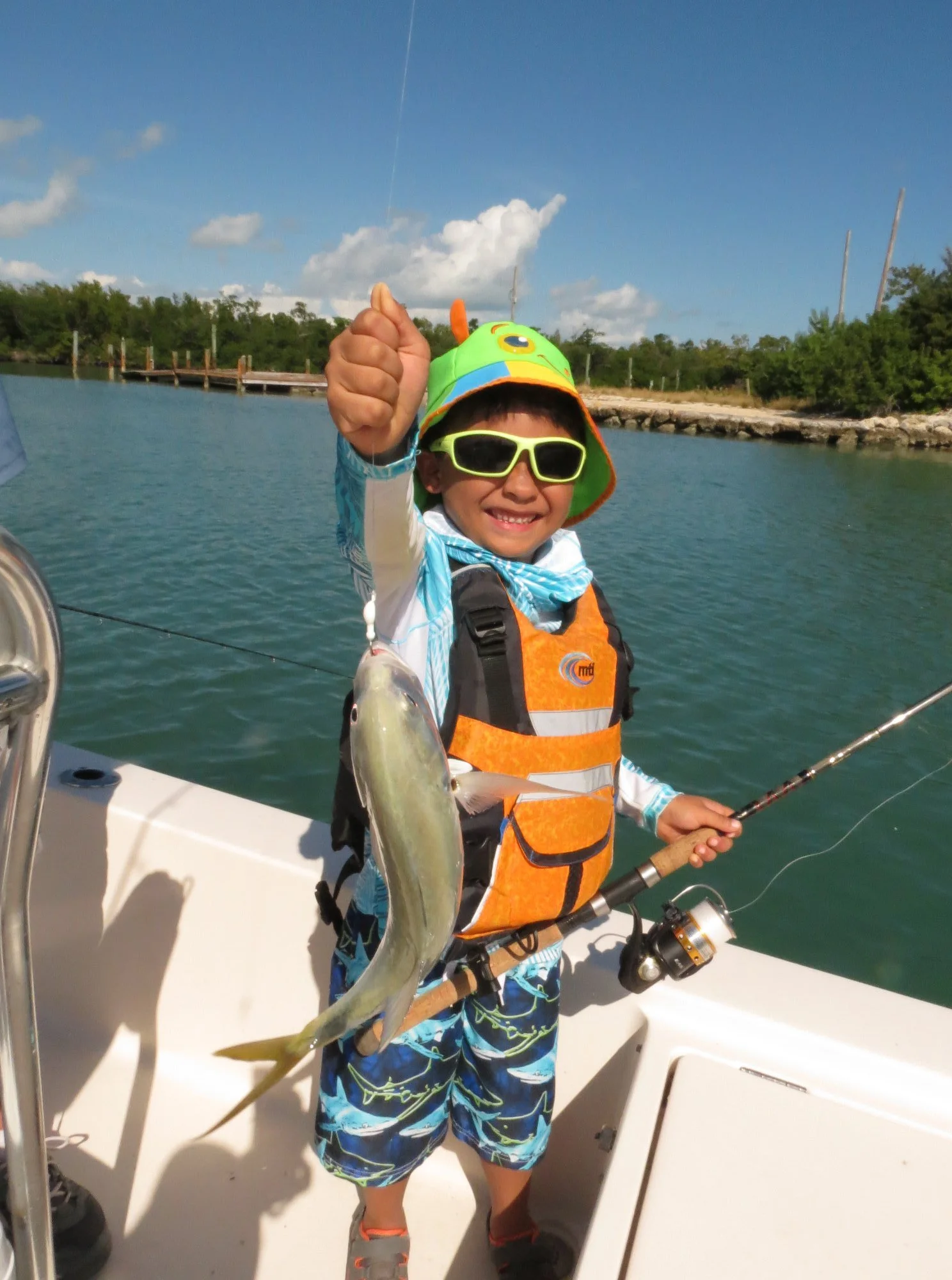 A young boy wearing sunglasses, a colorful hat, and an orange life jacket on a boat, holding up a fishing rod with a fish he caught, smiling at the camera.