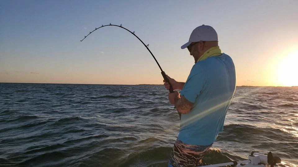A man fishing in the ocean at sunset, holding a fishing rod with a line cast into the water.