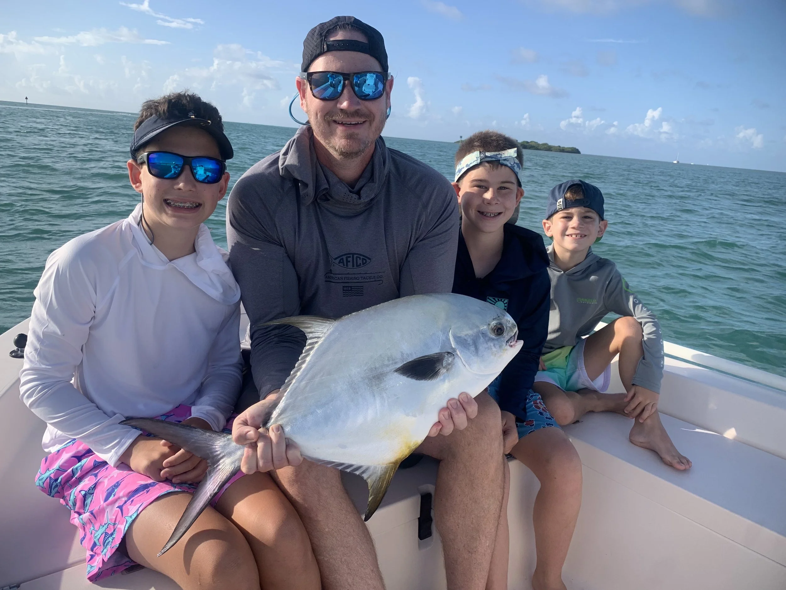 A man and three boys sitting on a boat holding a large fish, with the ocean and a partly cloudy sky in the background.