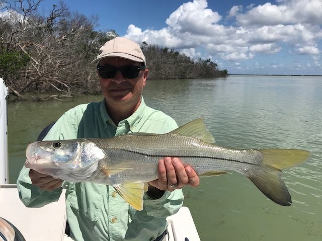 Person wearing sunglasses, a light-colored hat, and a light green shirt holding a large fish on a boat by a river or lake with trees and partly cloudy sky in the background.