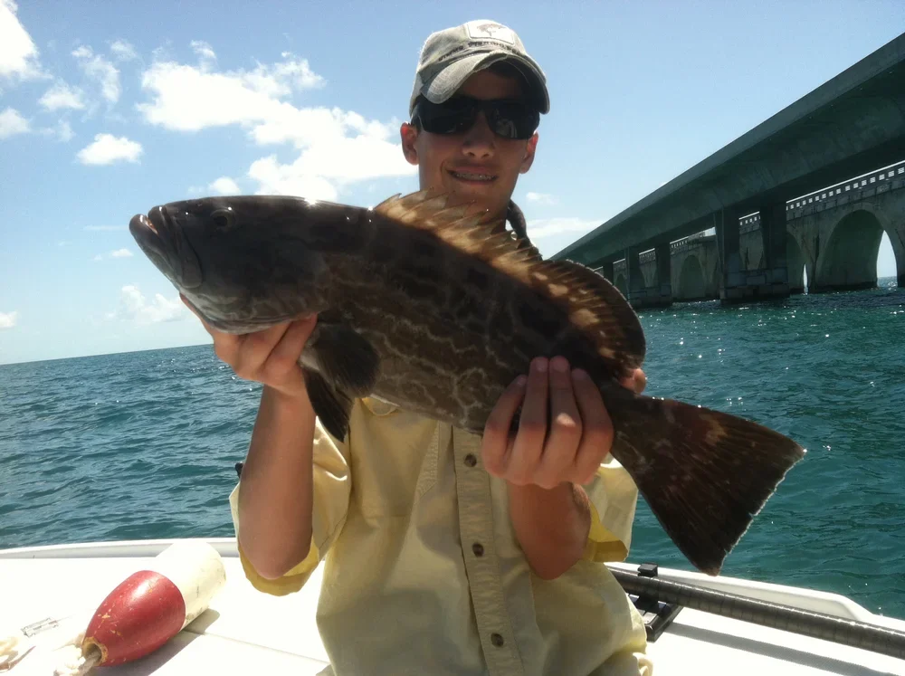A man wearing a hat and sunglasses on a boat holding a large fish over the water.