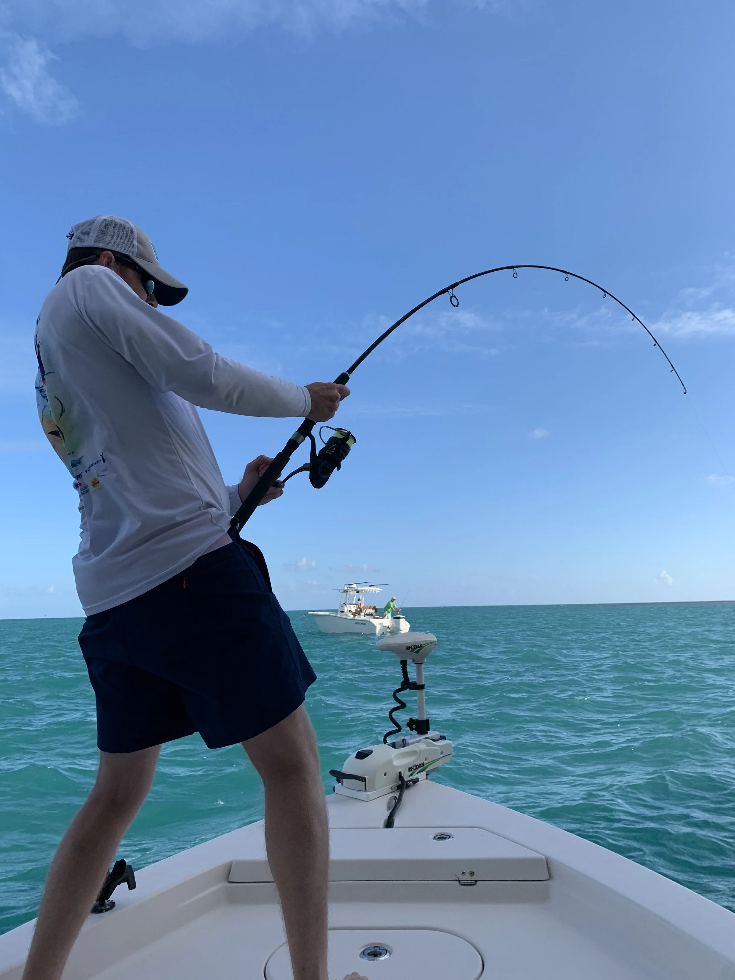 Person fishing from a boat on the water, with a boat in the distance under a clear blue sky.
