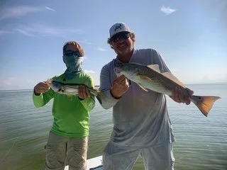 Two people holding fish on a beach with calm water and a clear sky.