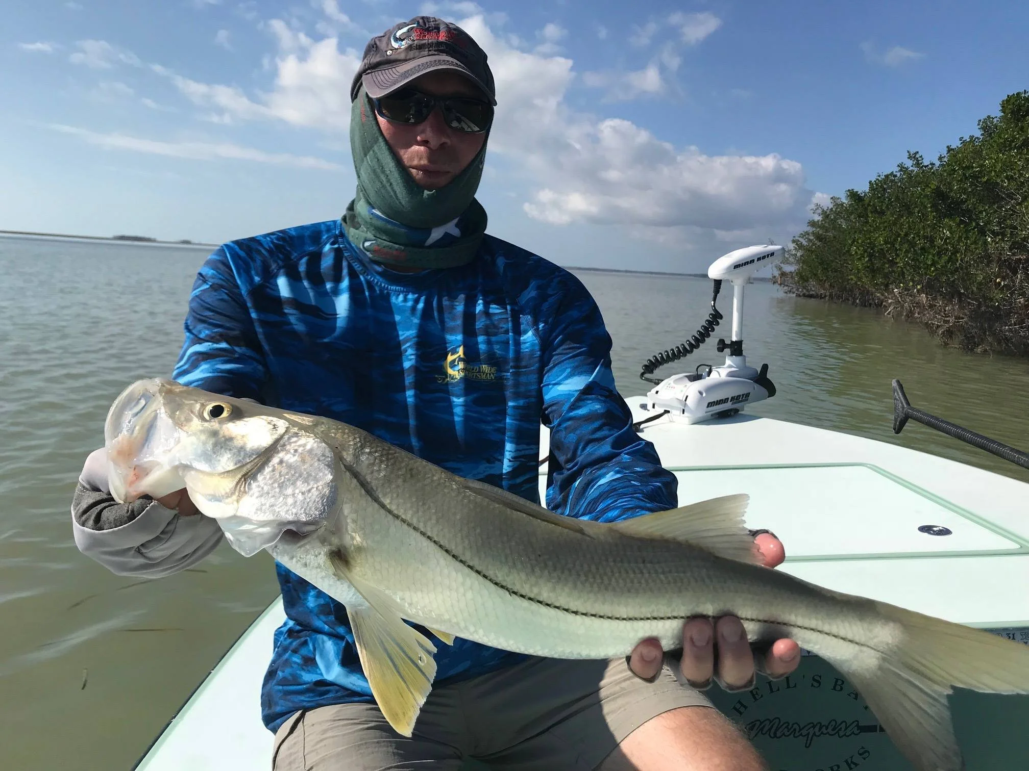 Man wearing a blue shirt, sunglasses, and a neck gaiter holding a large fish on a boat, with water and a shoreline with trees in the background.