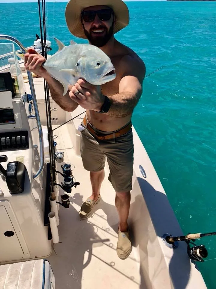 A man holding a fish on a boat in the water, smiling and wearing sunglasses and a wide-brim hat.
