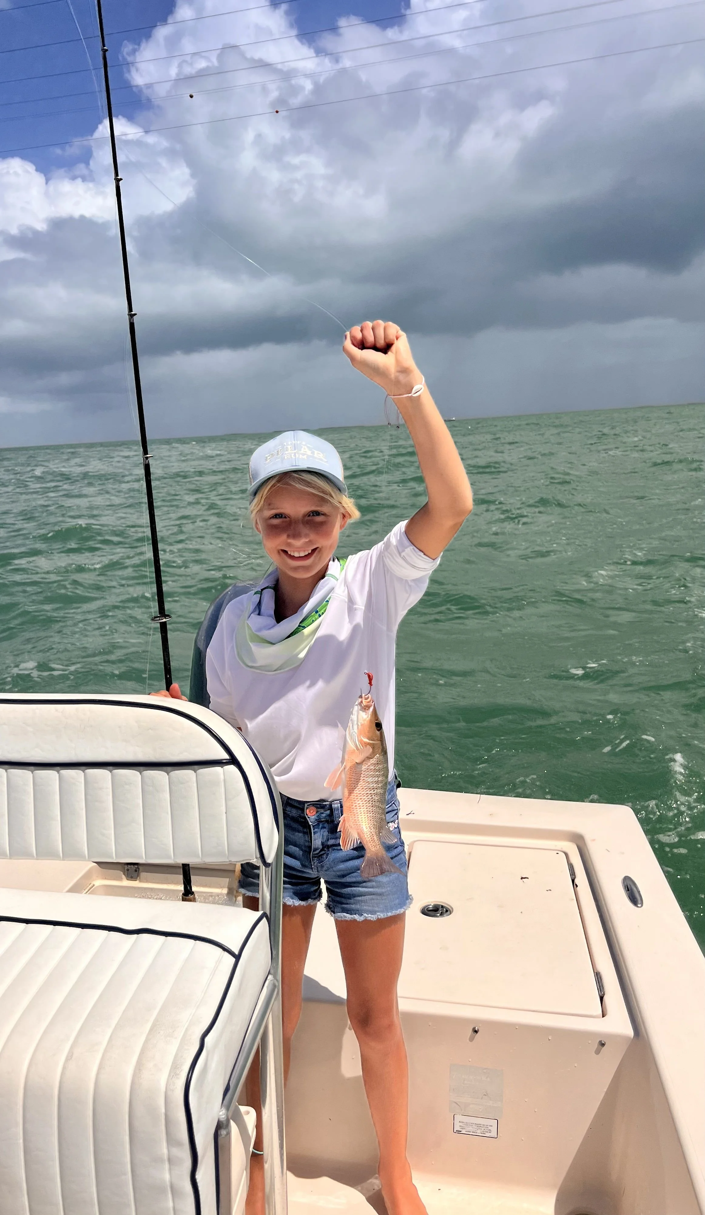 A girl on a boat holding a fishing rod with a fish she caught, smiling at the camera with body language of pride. Dark storm clouds are in the background over green water.