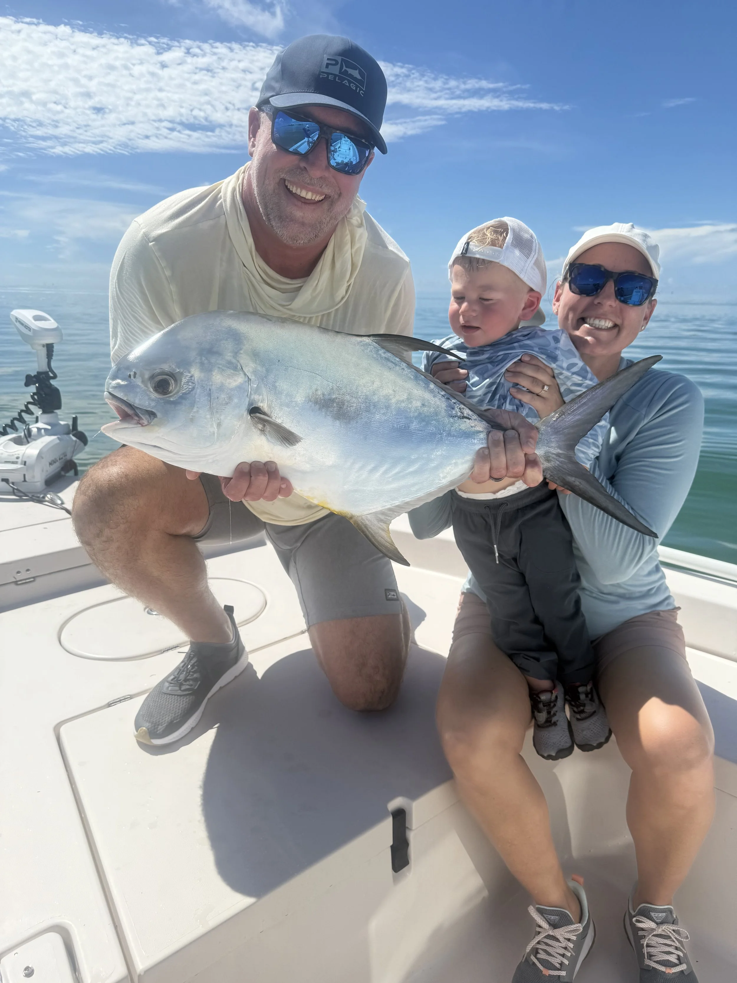 Family on a boat holding a large fish they caught, smiling at the camera, with blue sky and water in the background.