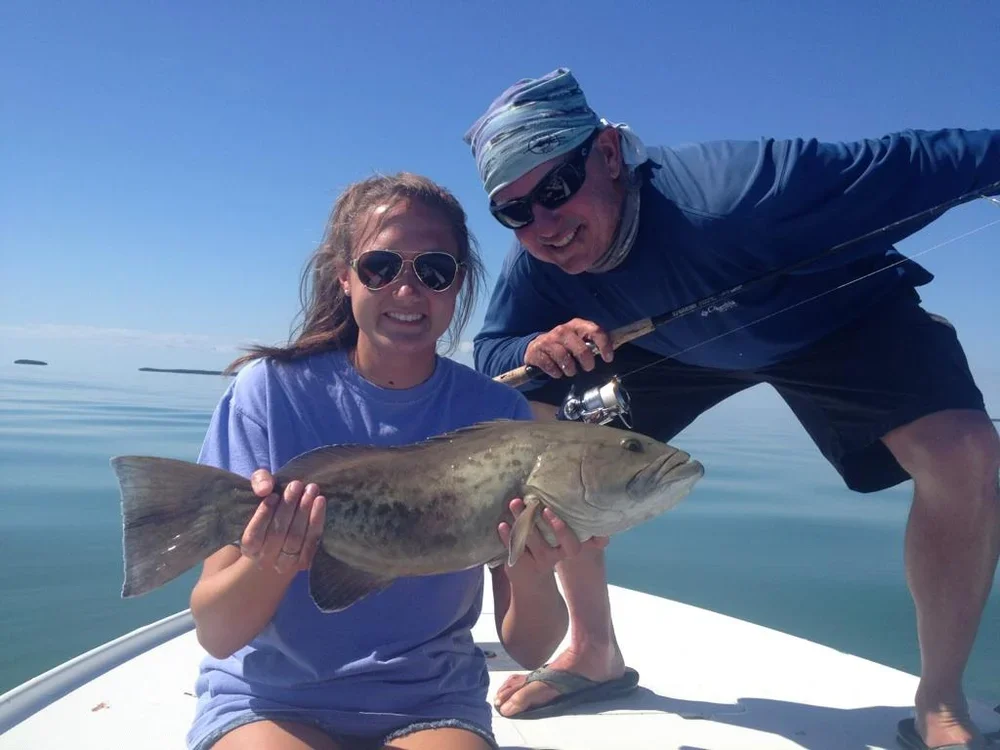 A woman holding a large fish on a boat with a man, both smiling with the ocean in the background.