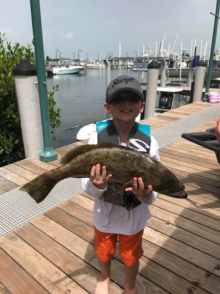 A young boy standing on a wooden dock holding a large fish he caught. He is wearing a gray cap, sunglasses, a white long-sleeve shirt, orange shorts, and a life jacket. In the background, there are boats docked in the marina, with water and cloudy sk