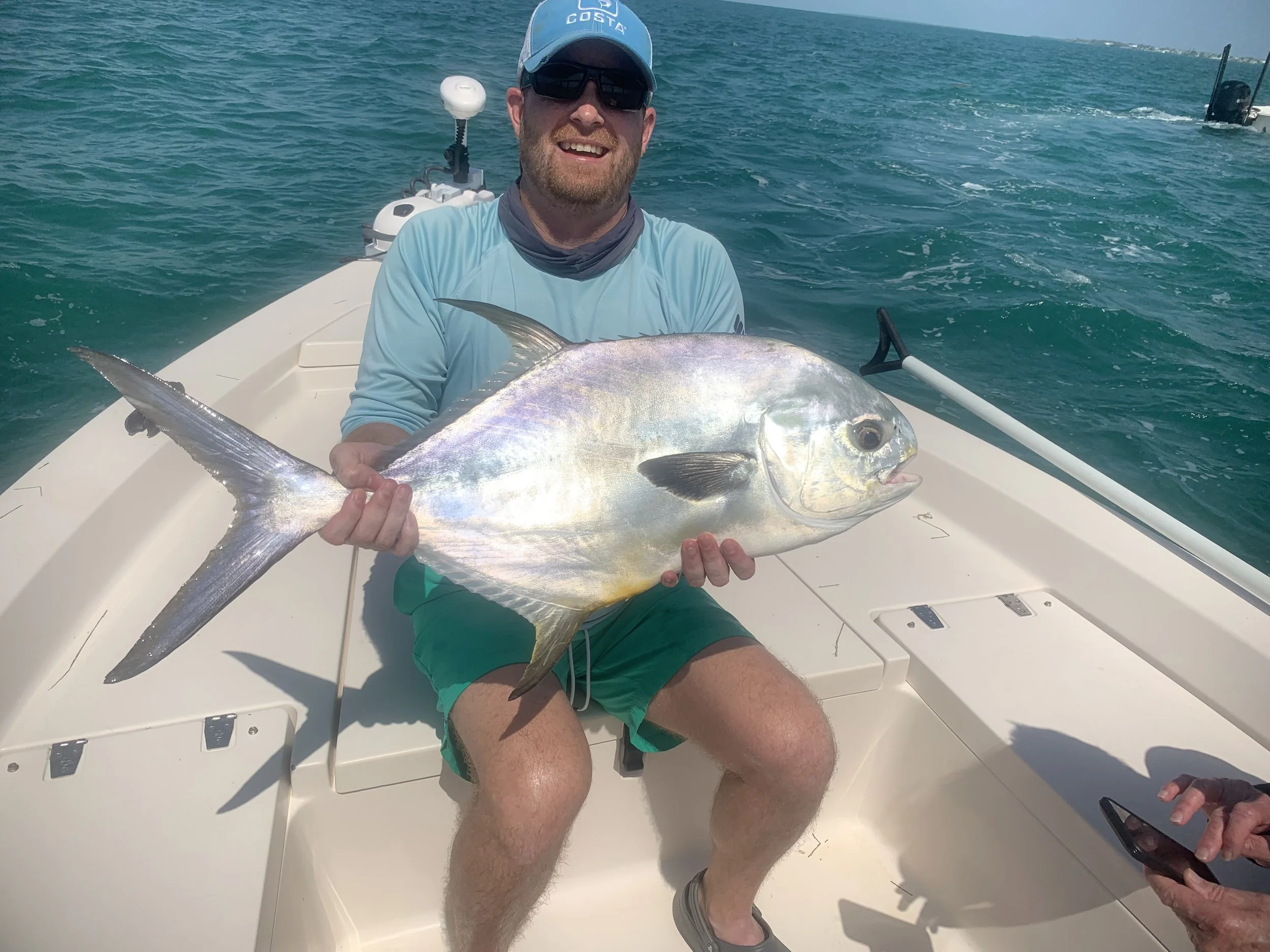 A man wearing sunglasses, a blue cap, and a light blue shirt sitting on a white boat, holding a large fish with silver and gold scales, on a blue ocean with more boats in the background.