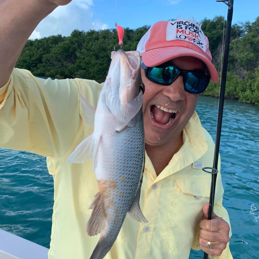 Man in yellow shirt, wearing sunglasses and a pink cap, holding a fishing rod and proudly displaying a caught fish on a boat with water and green hills in the background.