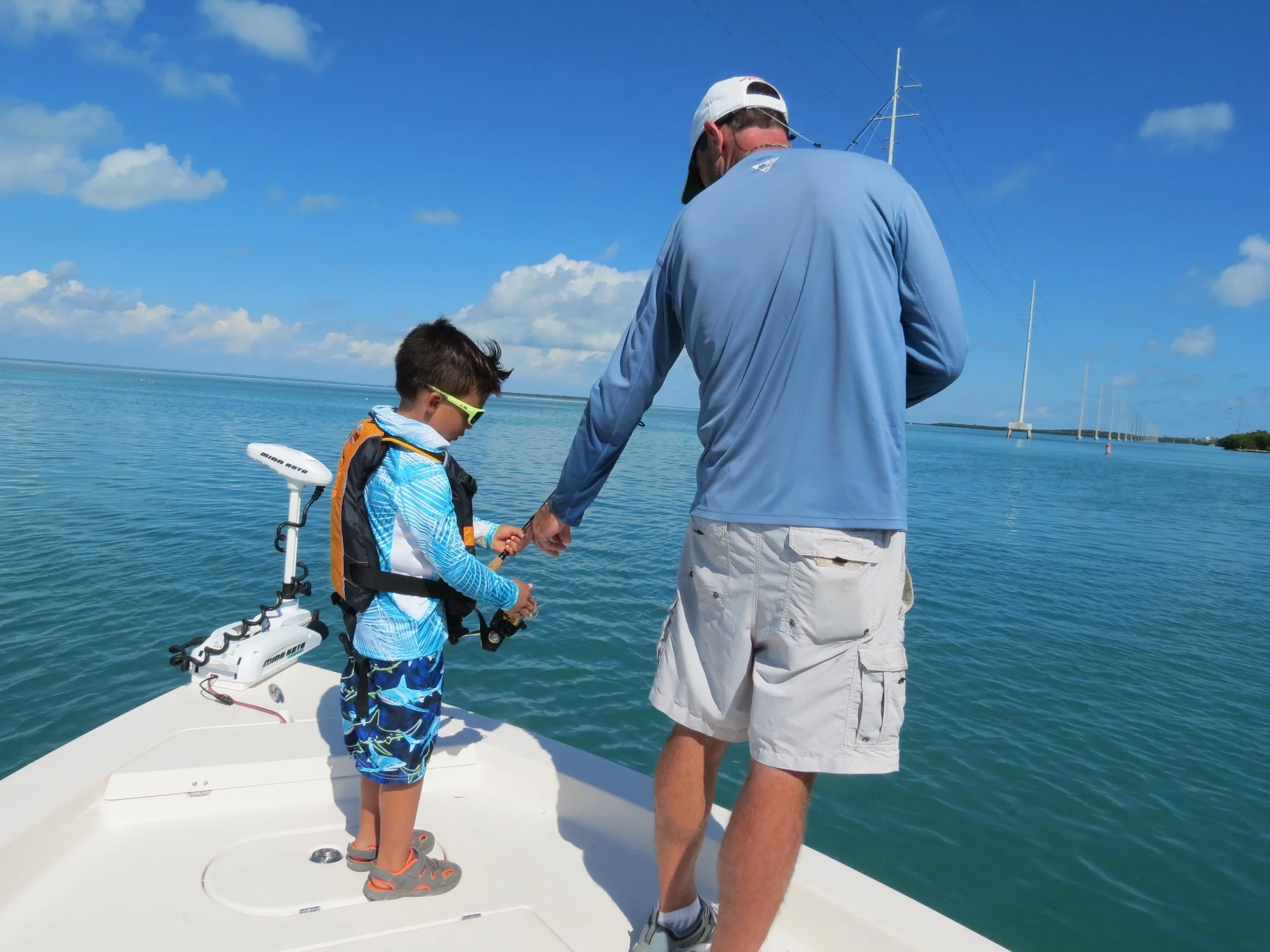 An adult and a young boy fishing together on a boat in a calm body of water on a sunny day, with sailboats in the distance.