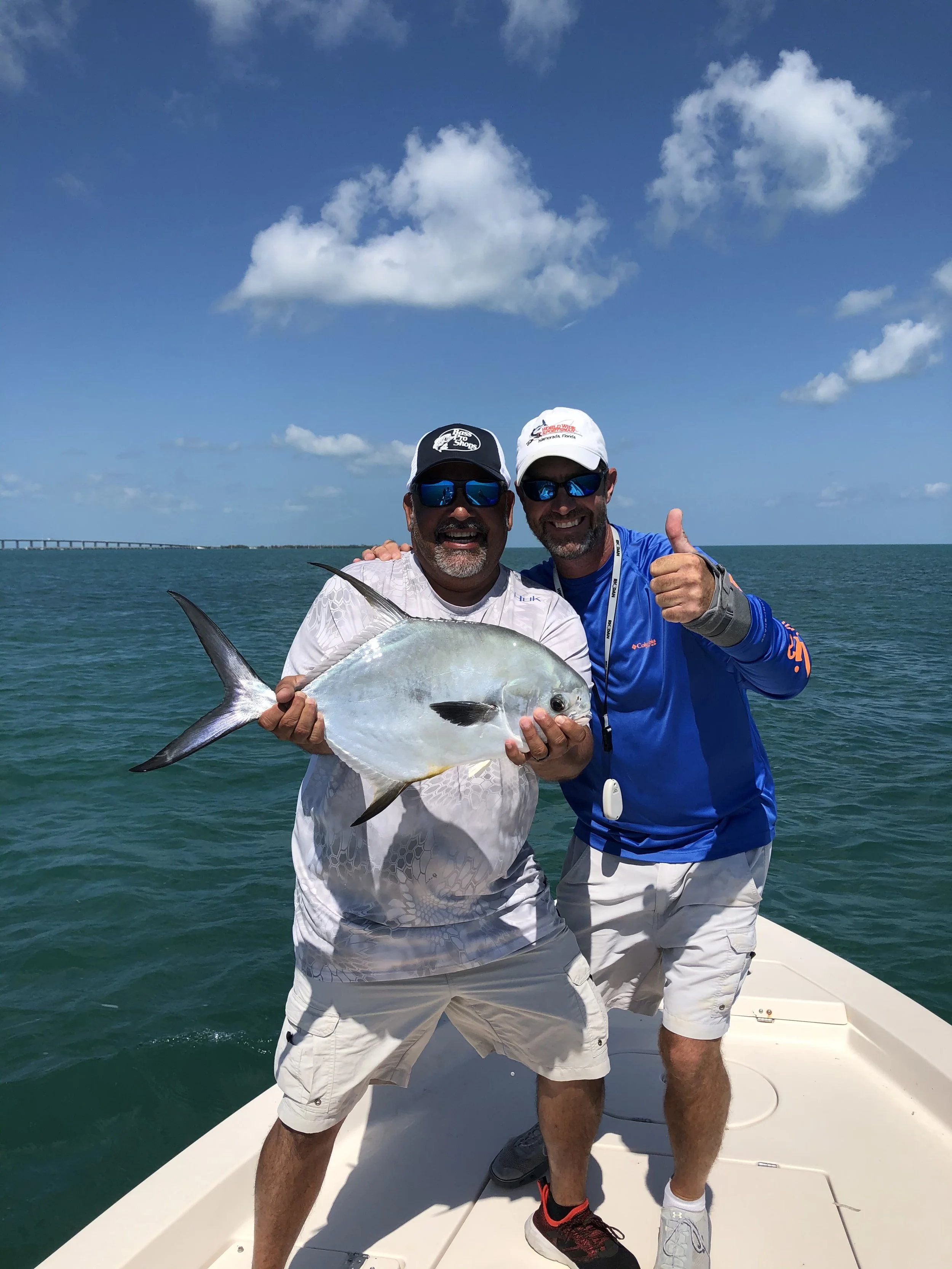 Two men on a boat holding a fish they caught, with the ocean and a bridge in the background.