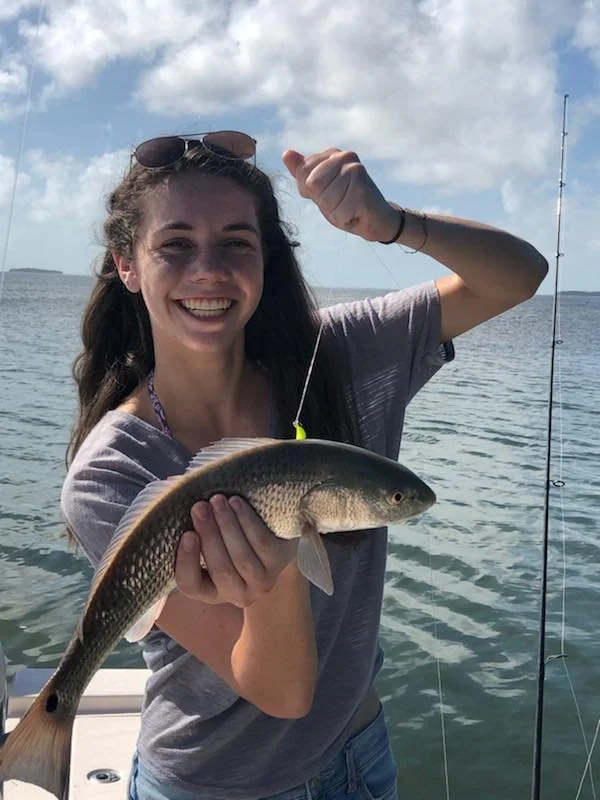 A young woman on a boat holding a fish she caught, with a fishing rod visible, smiling at the camera with sunglasses on her head, against a backdrop of water and a partly cloudy sky.