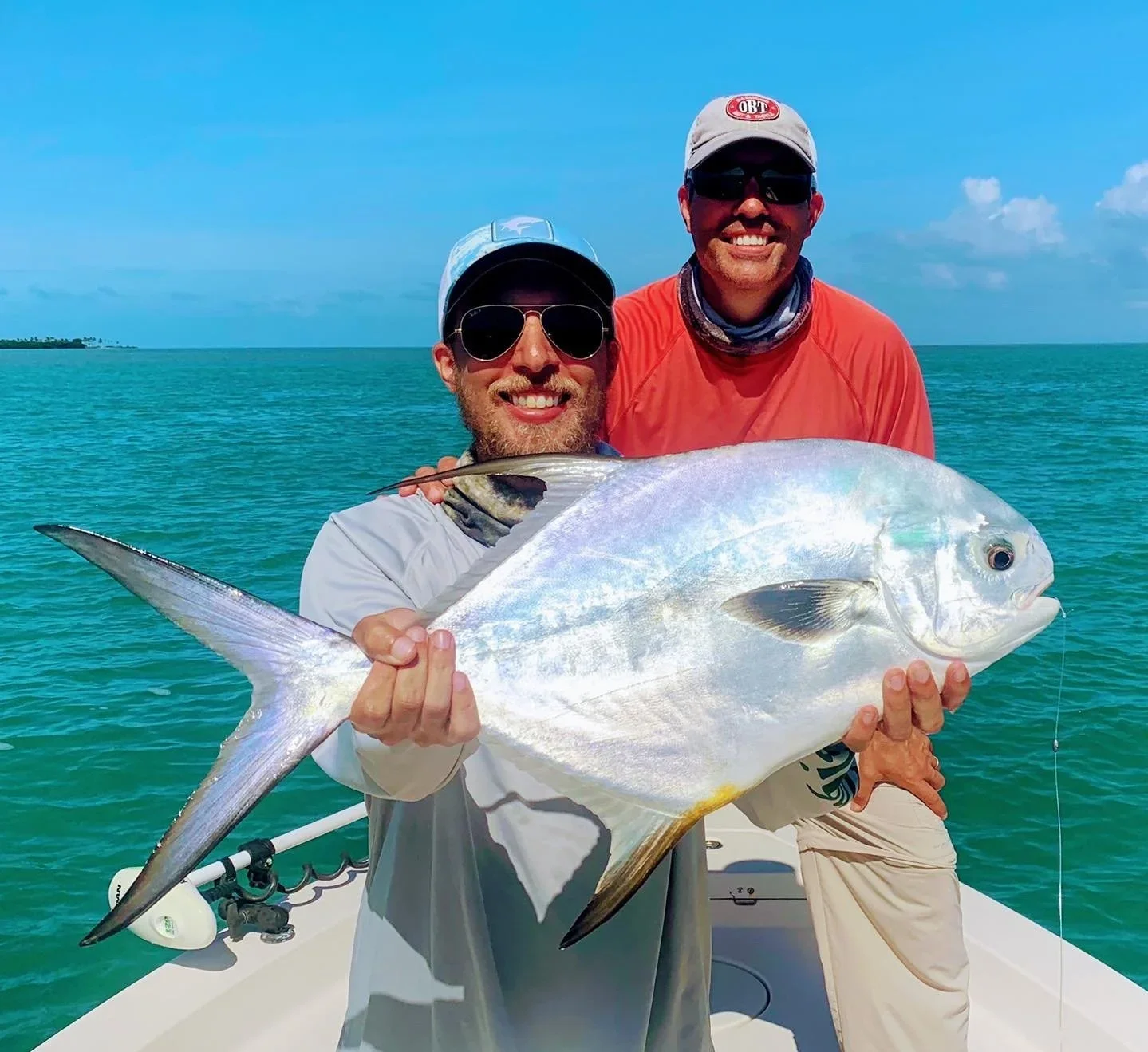 Two men on a boat holding a large, shiny fish with a silver body and a forked tail, with ocean and blue sky in the background.