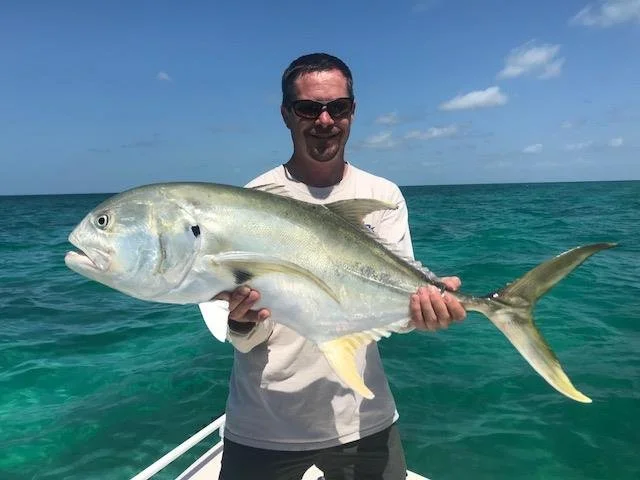 Man holding a large fish on a boat in the ocean, with blue sky and clouds in the background.