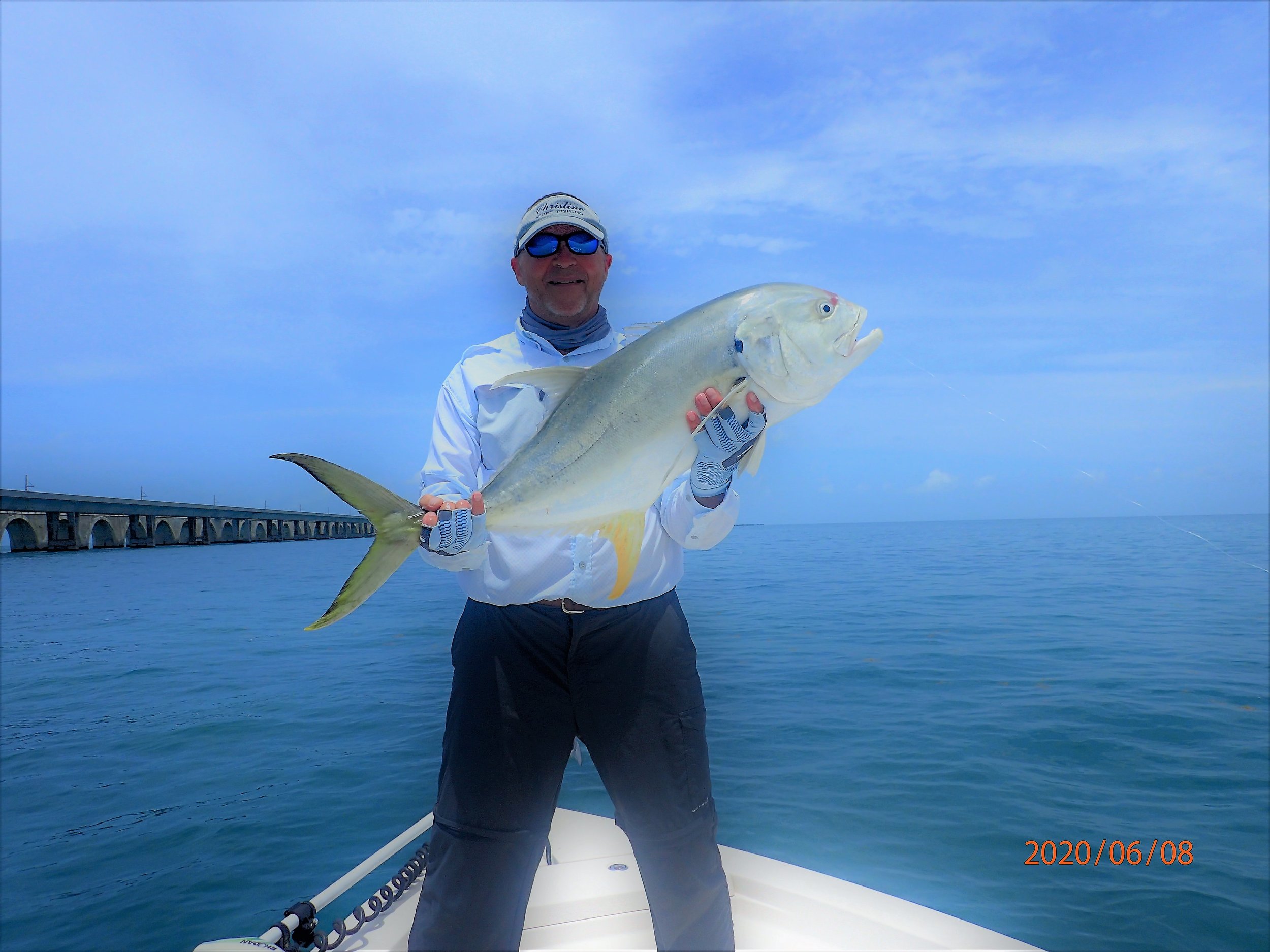 A man in fishing gear holding a large fish on a boat, with water and a bridge in the background on a cloudy day.