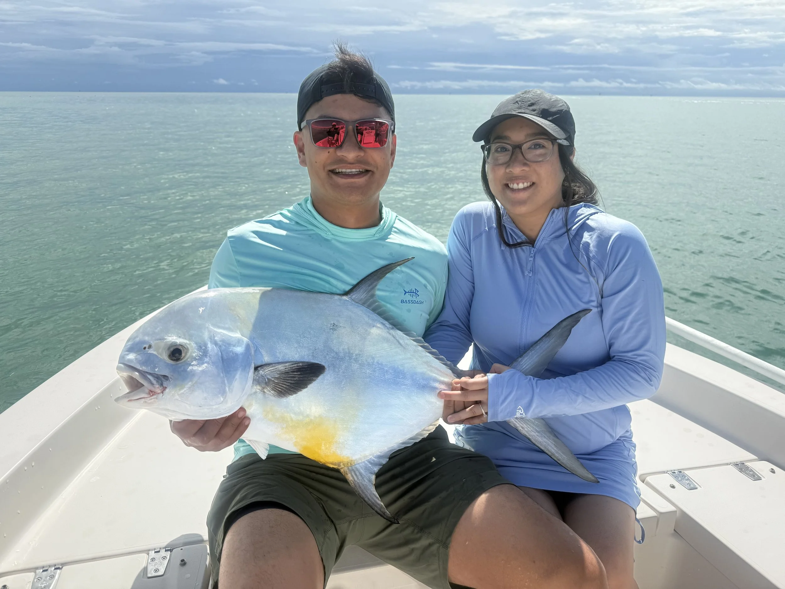 A man and a woman on a boat holding a large fish they caught, with a body of water and a cloudy sky in the background.