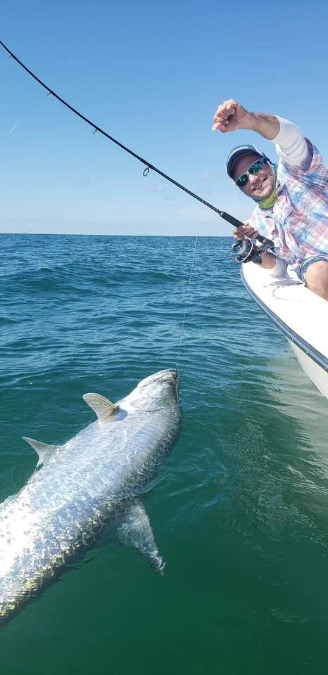 Man smiling on a boat holding a fishing rod with a caught large fish in the water near the boat.
