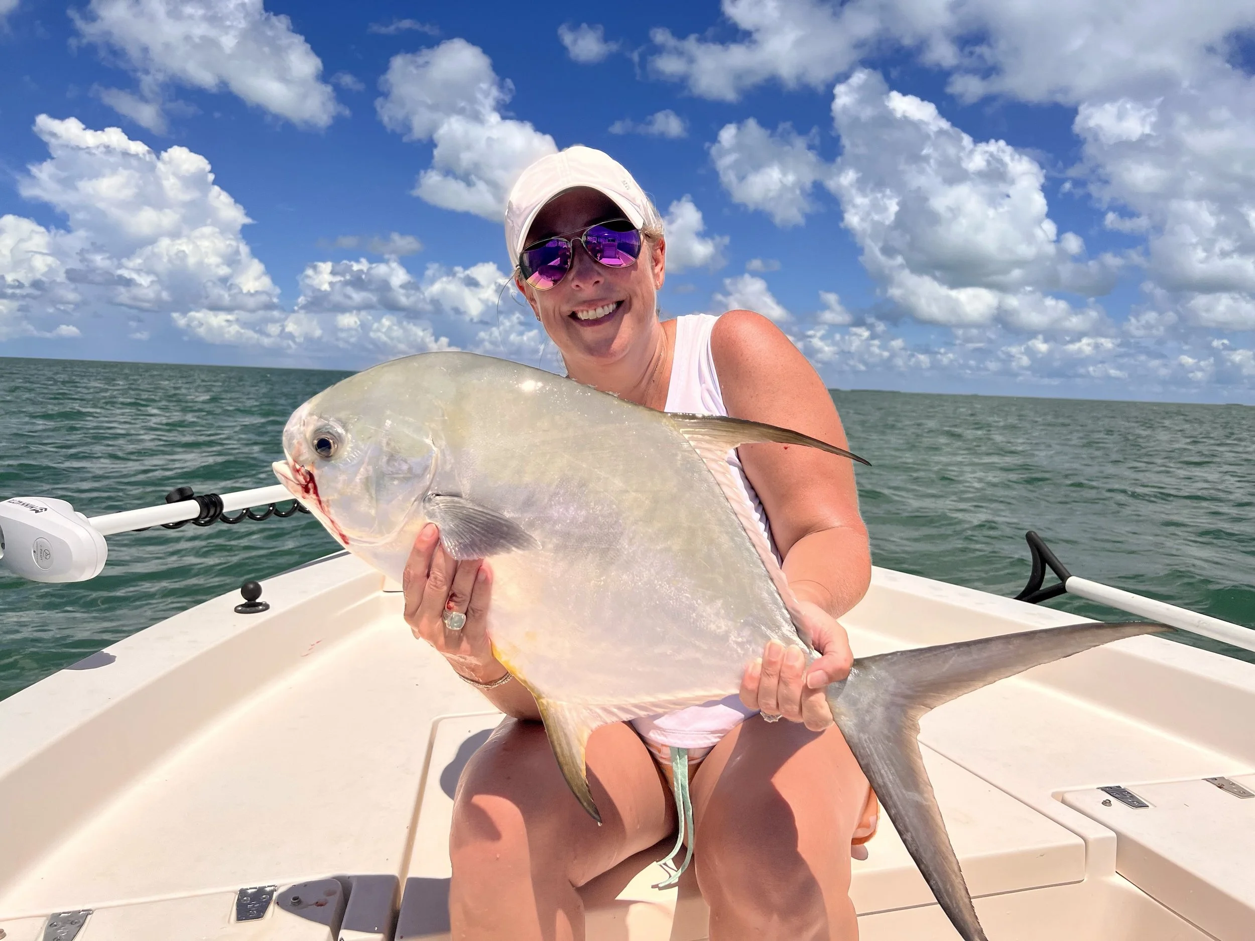 A woman wearing sunglasses and a white cap on a boat, holding a large fish against a backdrop of blue sky with clouds and ocean water.