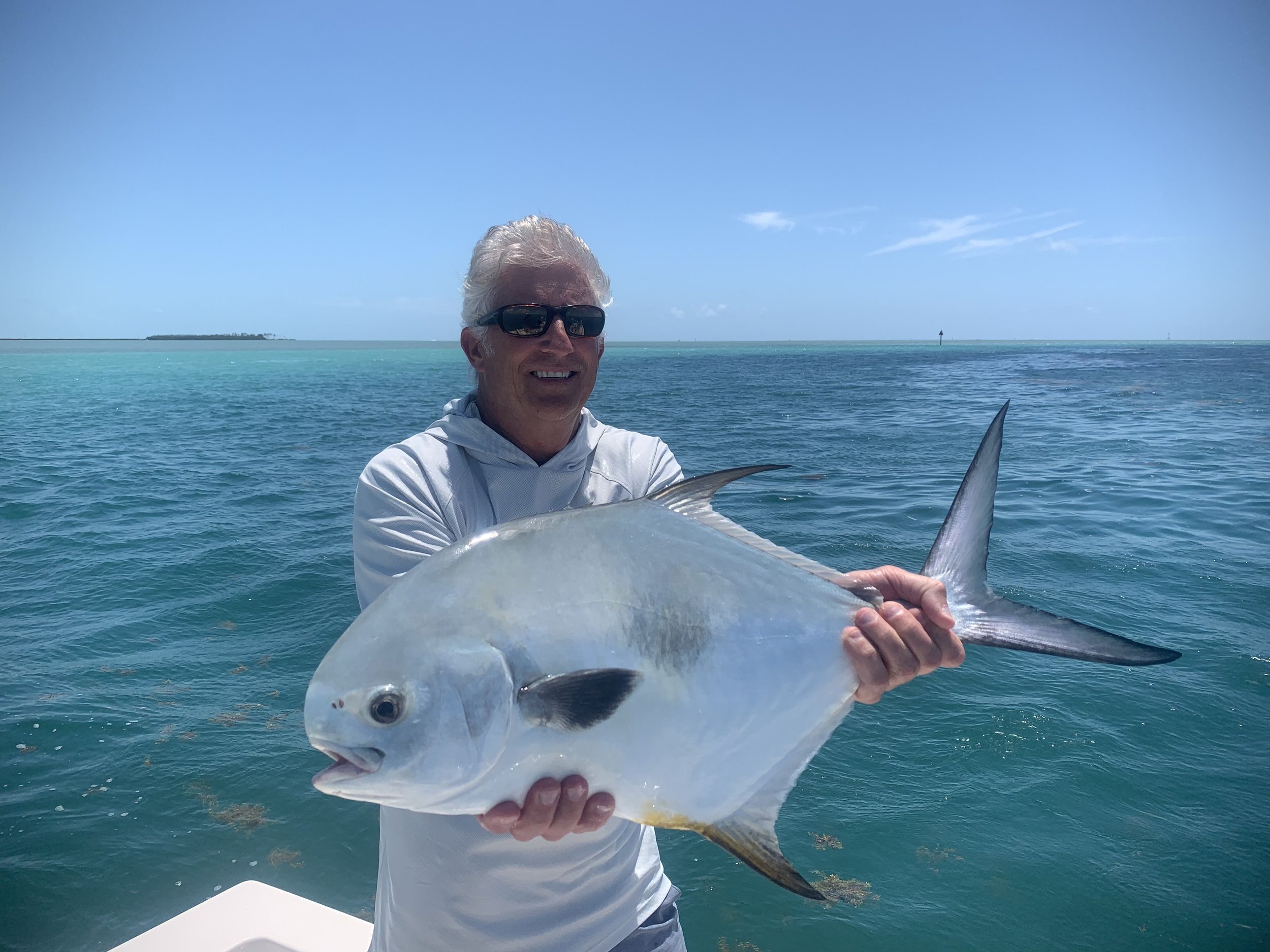 A man with gray hair and sunglasses holding a large fish on a boat, with the ocean and a distant landmass in the background.