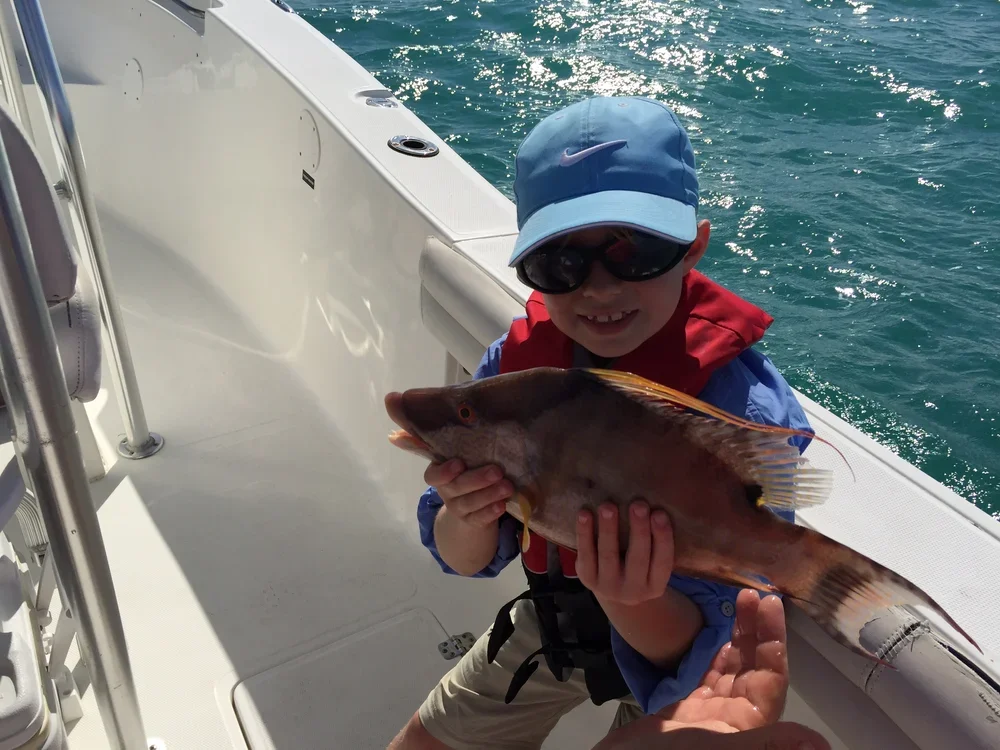 Young boy wearing a cap, sunglasses, and a life jacket on a boat, holding a large fish he caught.