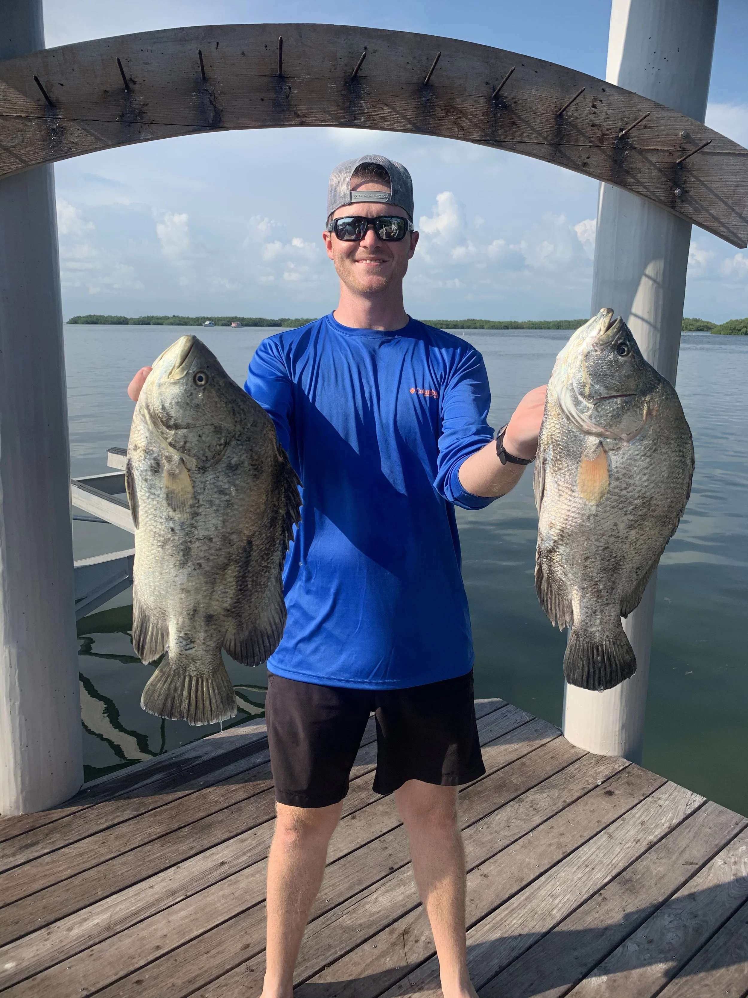 A man wearing sunglasses, a cap, a blue long-sleeve shirt, and black shorts standing on a wooden dock holding two large fish, one in each hand, with water and a cloudy sky in the background.