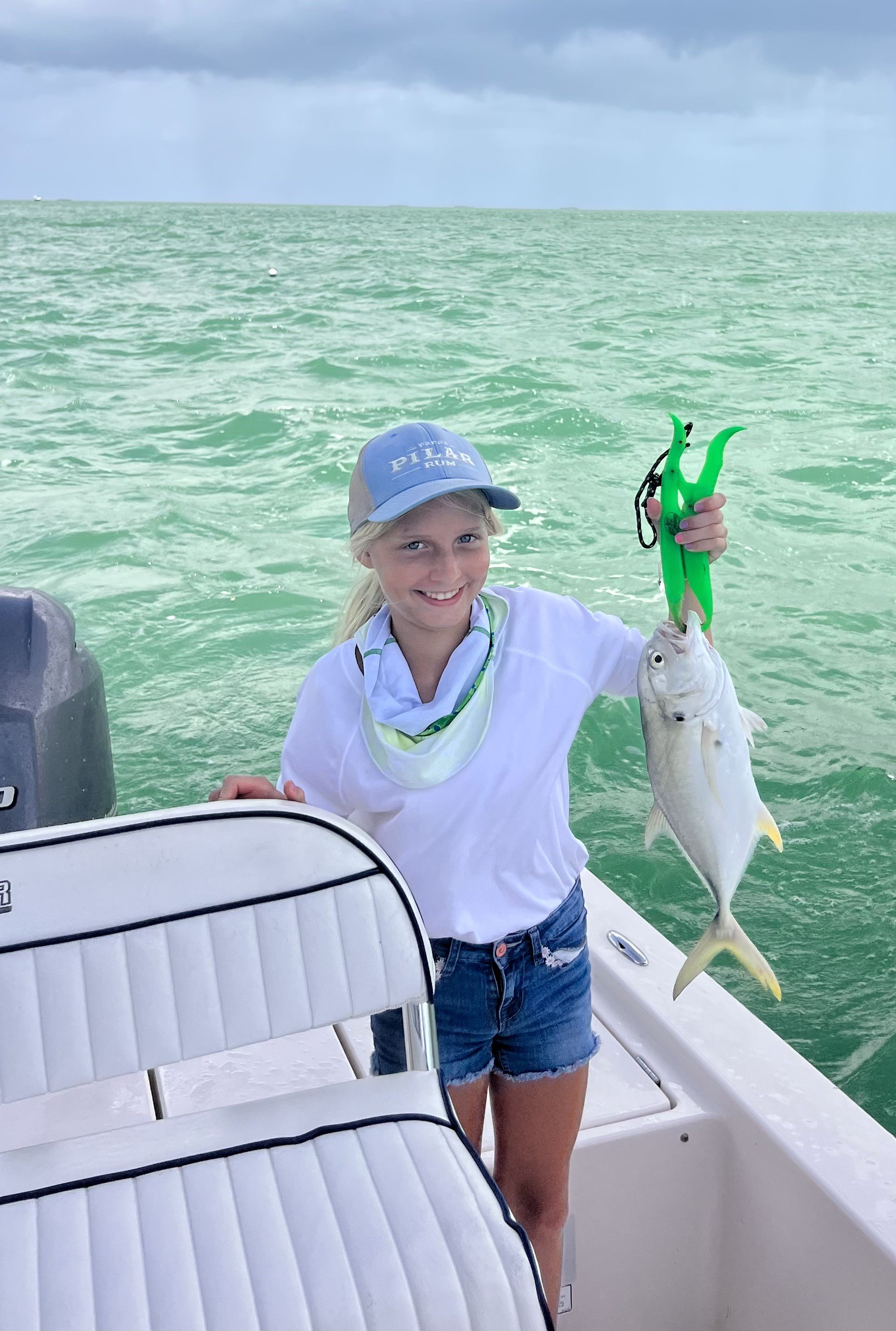 A young girl on a boat holding a fish she caught with a fish gripper. The water is green and the sky is cloudy.