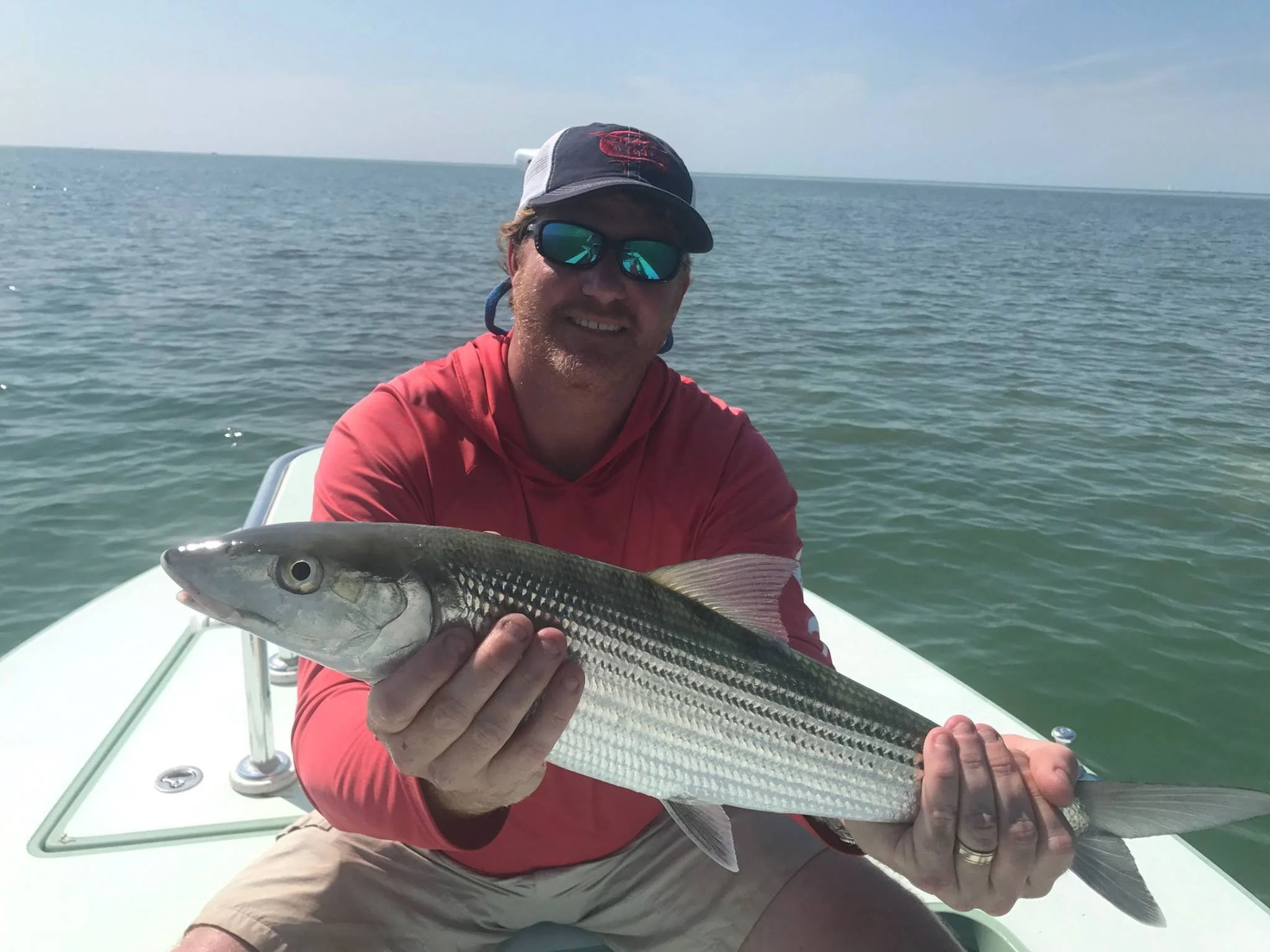 Man on boat holding a striped fish, wearing a red shirt, sunglasses, and a cap, with open water and sky in the background.