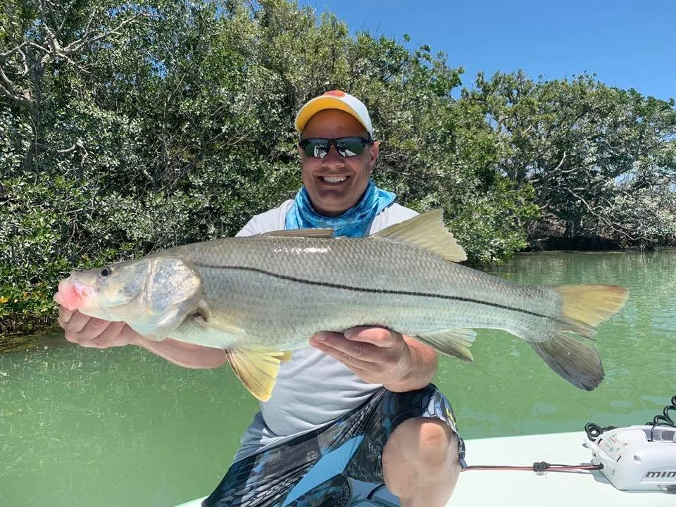 Man wearing sunglasses, a cap, and a blue neck gaiter, smiling and holding a large fish with both hands on a boat in a green water body with trees in the background.