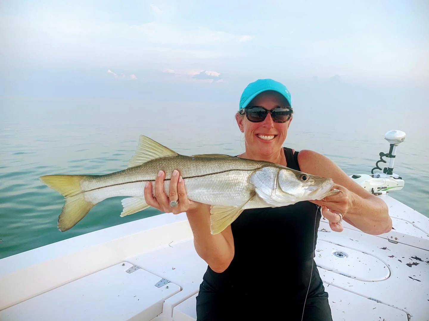Woman on a boat holding a large fish, smiling, wearing sunglasses and a blue cap, with water and sky in the background.