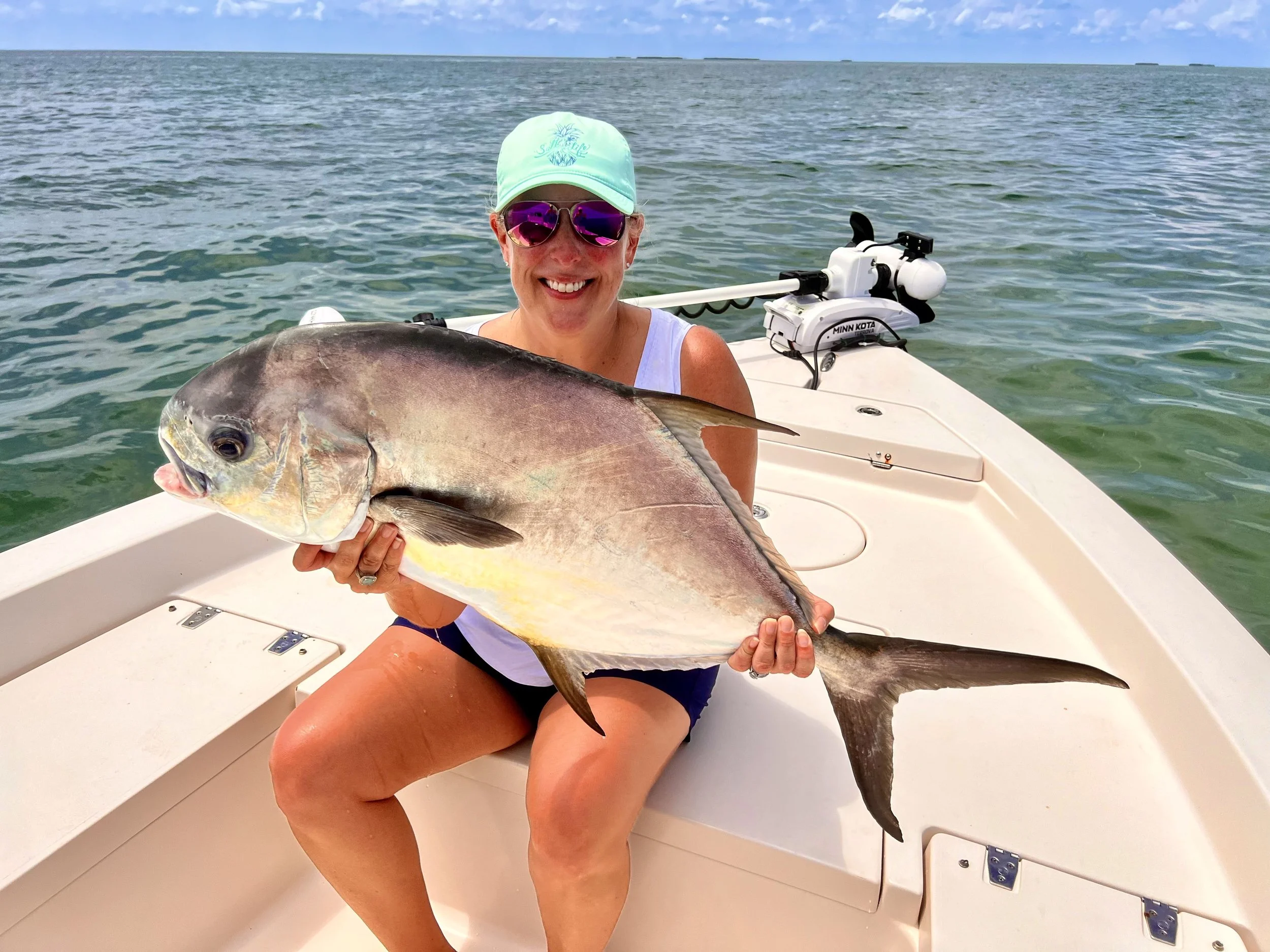 Woman sitting on a boat holding a large fish, wearing sunglasses, a cap, and casual clothing, with water and open sky in the background.