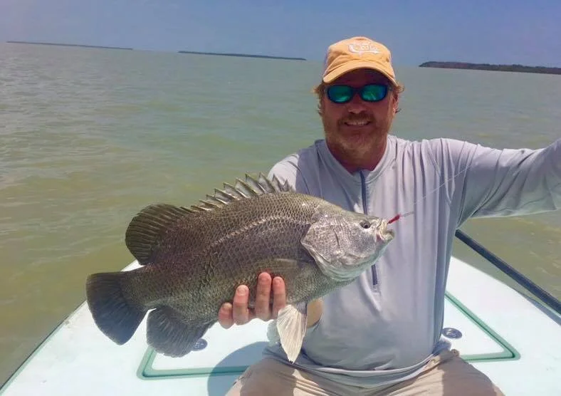Man on a boat holding a large fish in his hands, smiling, wearing a beige cap and sunglasses with a body of water in the background.