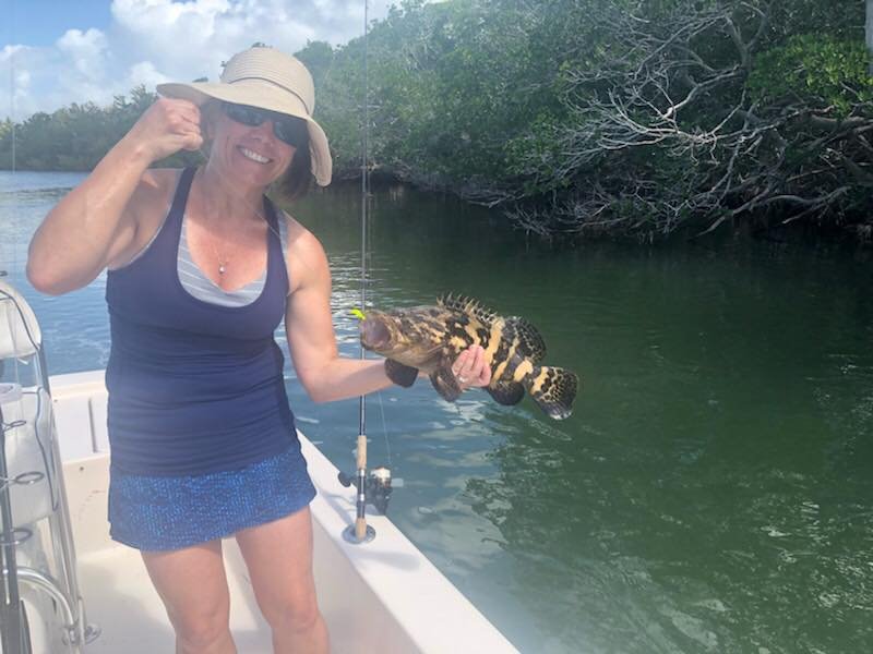 Woman wearing a sunhat and sunglasses holding a fish she caught on a boat with a fishing rod, with water and trees in the background.
