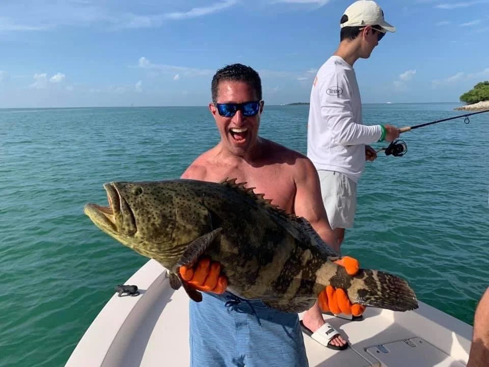 A man smiling and wearing sunglasses is holding a large fish on a boat during a fishing trip, with another person fishing in the background on the water.