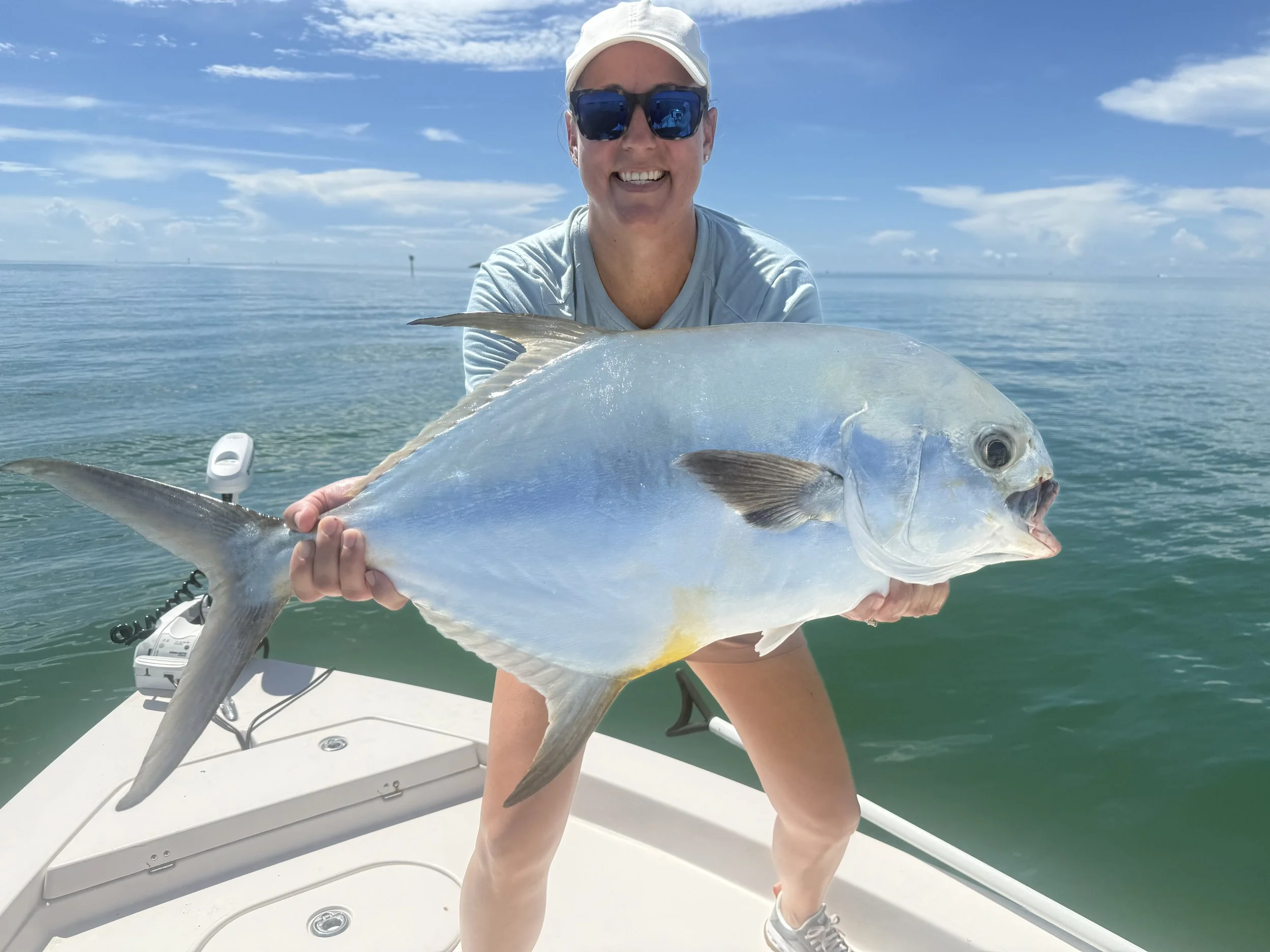Woman in sunglasses and a hat holding a large fish on a boat in the water, with a blue sky and clouds in the background.