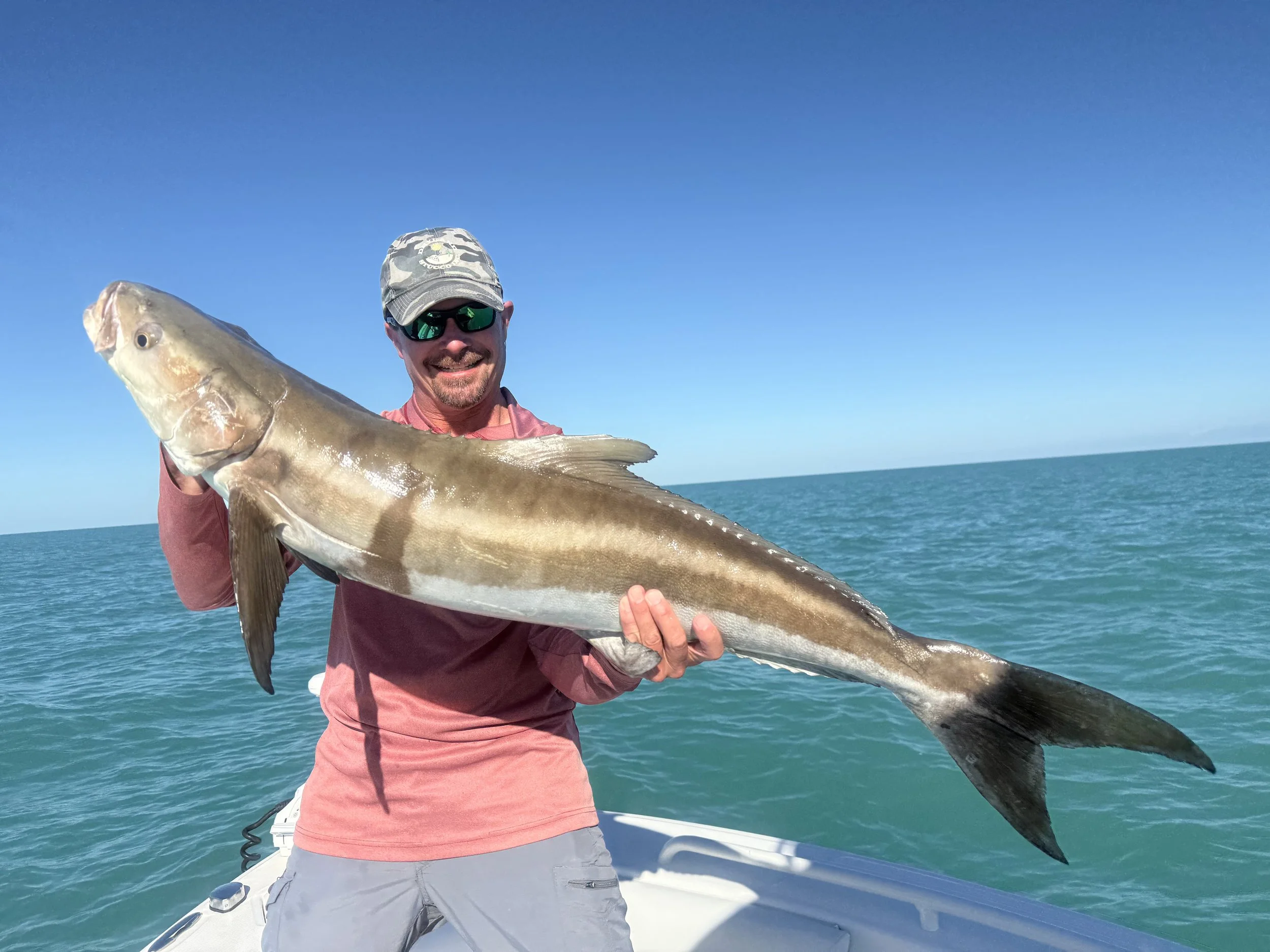 Man in sunglasses and hat holding large fish on boat in ocean under blue sky.