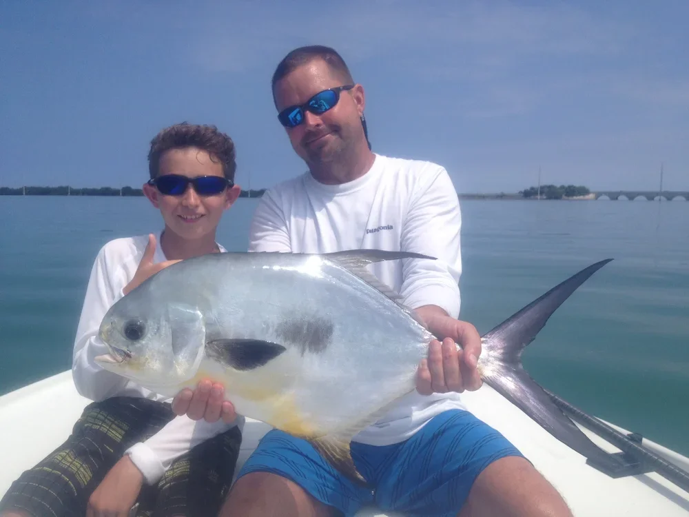 Two people on a boat holding a permit, with water and blue sky in the background.