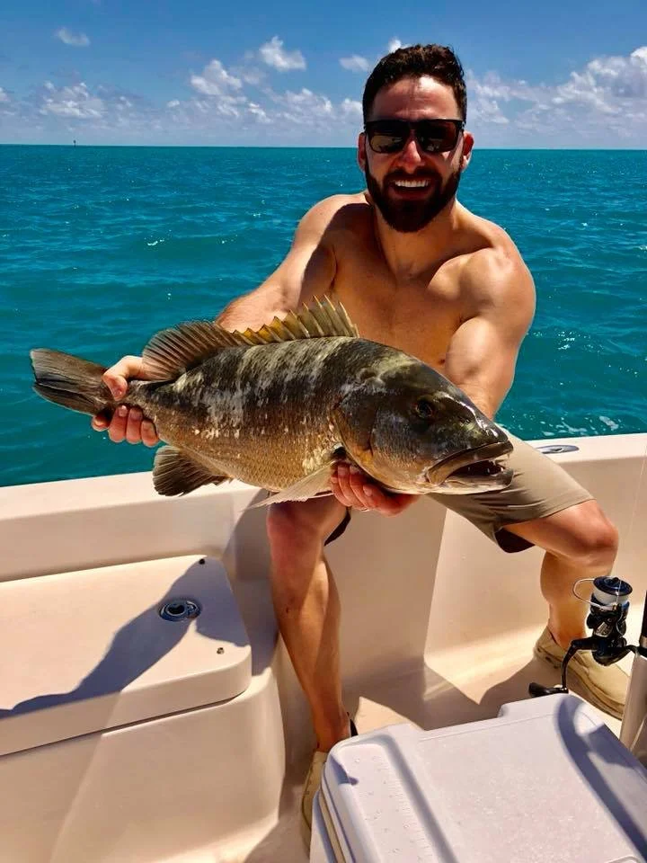 Man in sunglasses smiling while holding a large fish on a boat in the ocean under a partly cloudy sky.