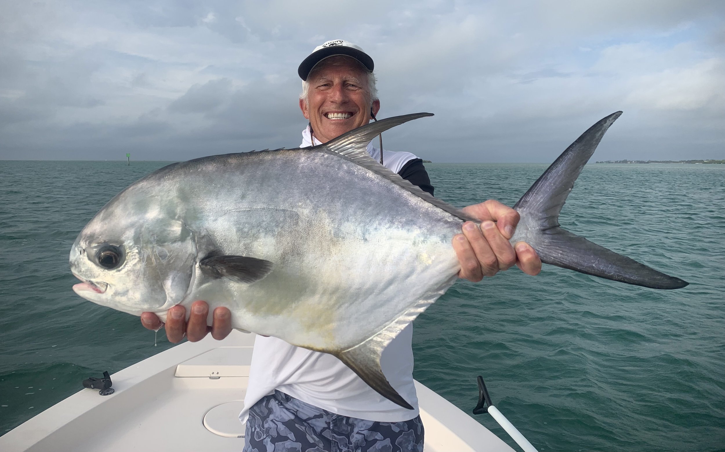 Man on a boat holding a large fish with a smiling expression, ocean and cloudy sky in the background.