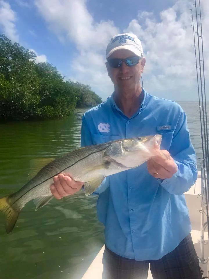 A man wearing a blue shirt, sunglasses, and a white cap is holding a large fish aboard a boat. The background shows water, some green trees, and a partly cloudy sky.