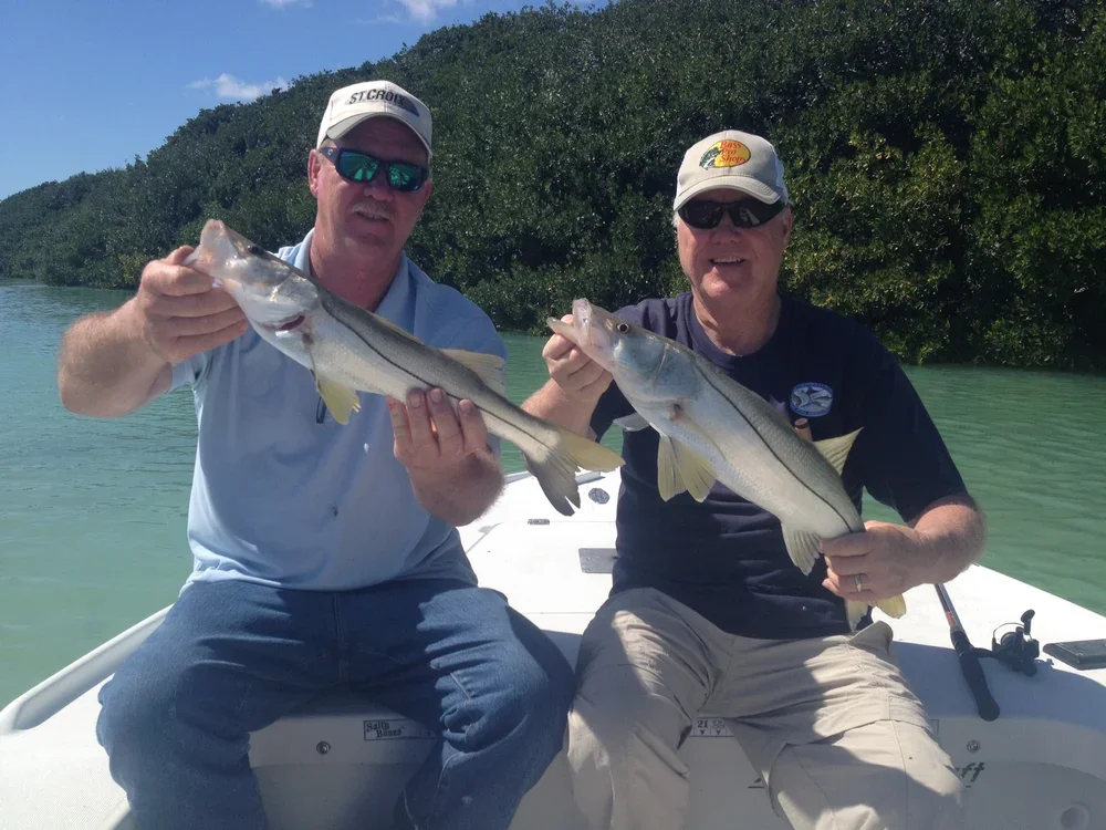 Two men sitting on a boat holding large fish, with water and greenery in the background.
