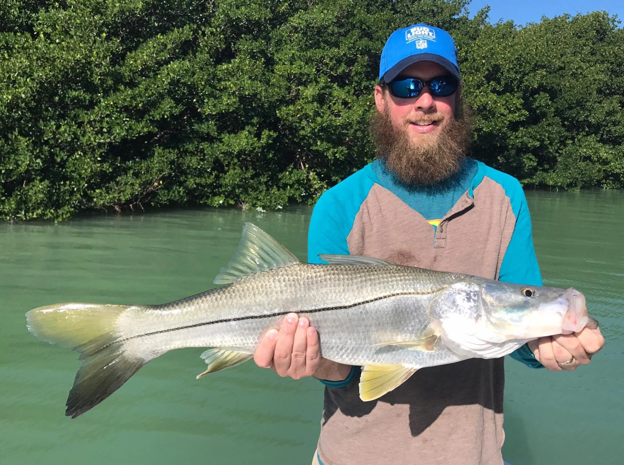 A man with a beard and sunglasses holding a large fish on a boat or near water, with lush green trees in the background.