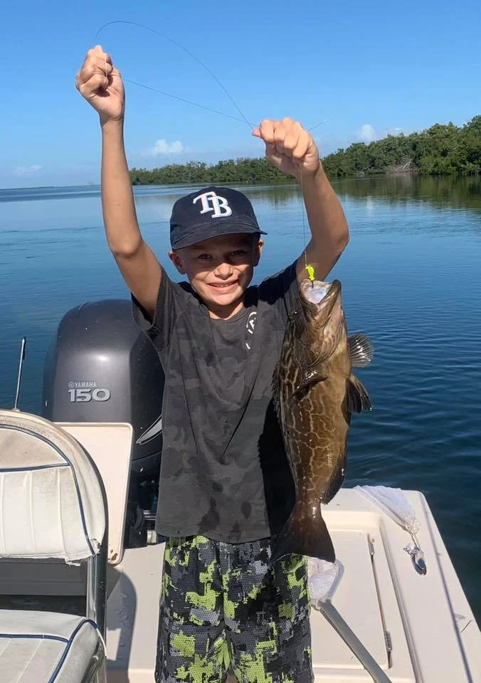 A young boy wearing a black Tampa Bay baseball cap and camouflage shorts on a boat, holding a large fish with both hands, smiling with his arms raised, with a calm body of water and green trees in the background.