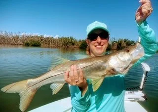 Person in a light blue jacket and hat holding a large fish on a boat, with water and reeds in the background.