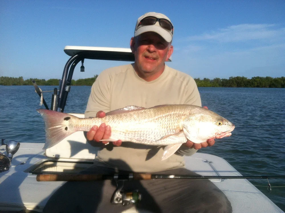 Man on a boat holding a large fish, with water and trees in the background, wearing a beige shirt, cap, and sunglasses.