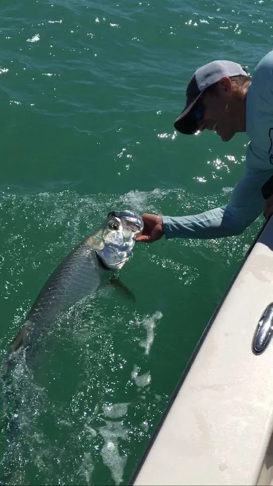 A person on a boat holding a large fish with both hands, near the boat's edge, surrounded by green water.