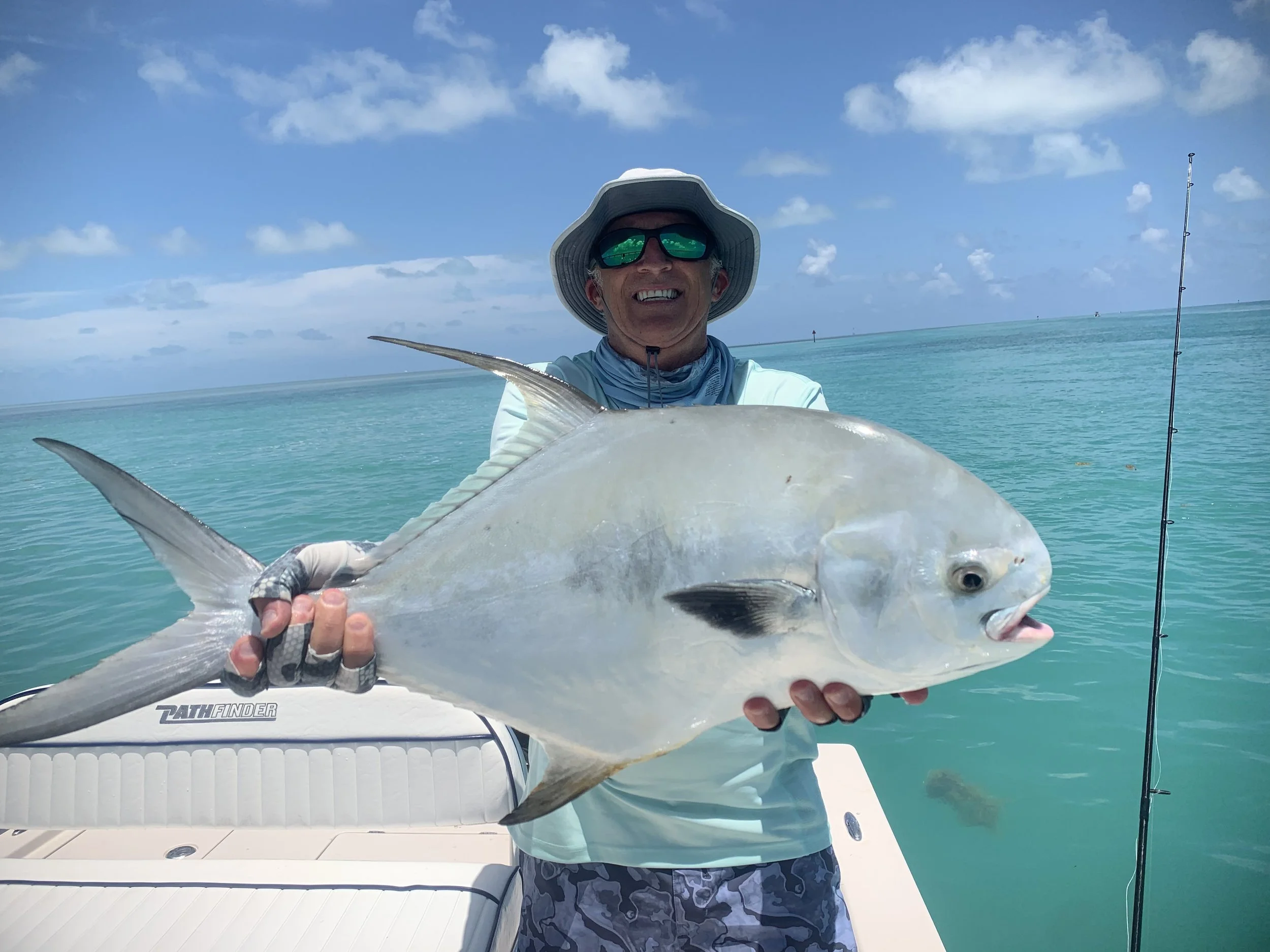 Man smiling while holding a large fish on a boat in turquoise ocean with a partly cloudy sky.