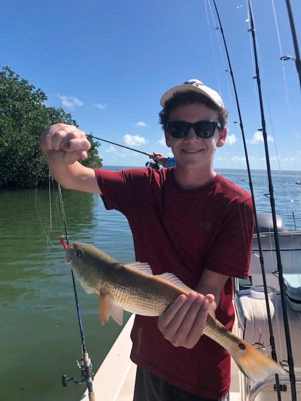 A young man wearing sunglasses and a white cap, holding a fishing rod, is on a boat showing off a caught fish with a broad smile. The fish is medium-sized with a tan body and darker fins. The background features a calm body of water, green trees, and